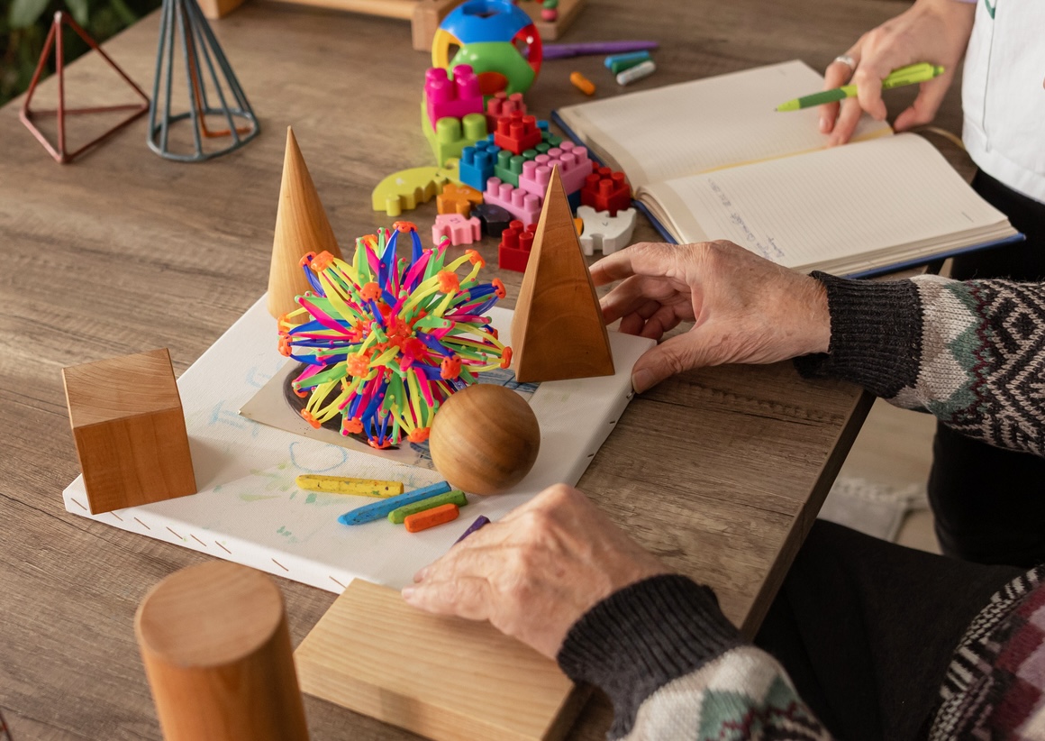 Elderly person arranging wooden geometric shapes and colorful toys on a table with an open notebook nearby.