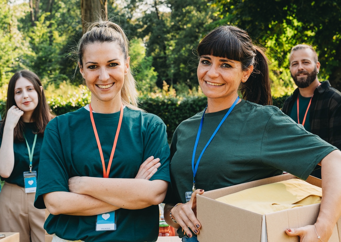 Four smiling volunteers outdoors wearing green shirts and badges, one holding a box of supplies.