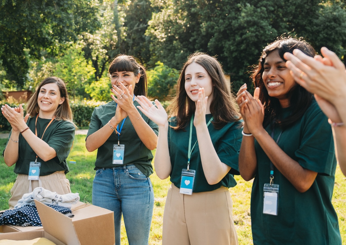 Four diverse young volunteers in green shirts smiling and clapping outdoors near donation boxes.