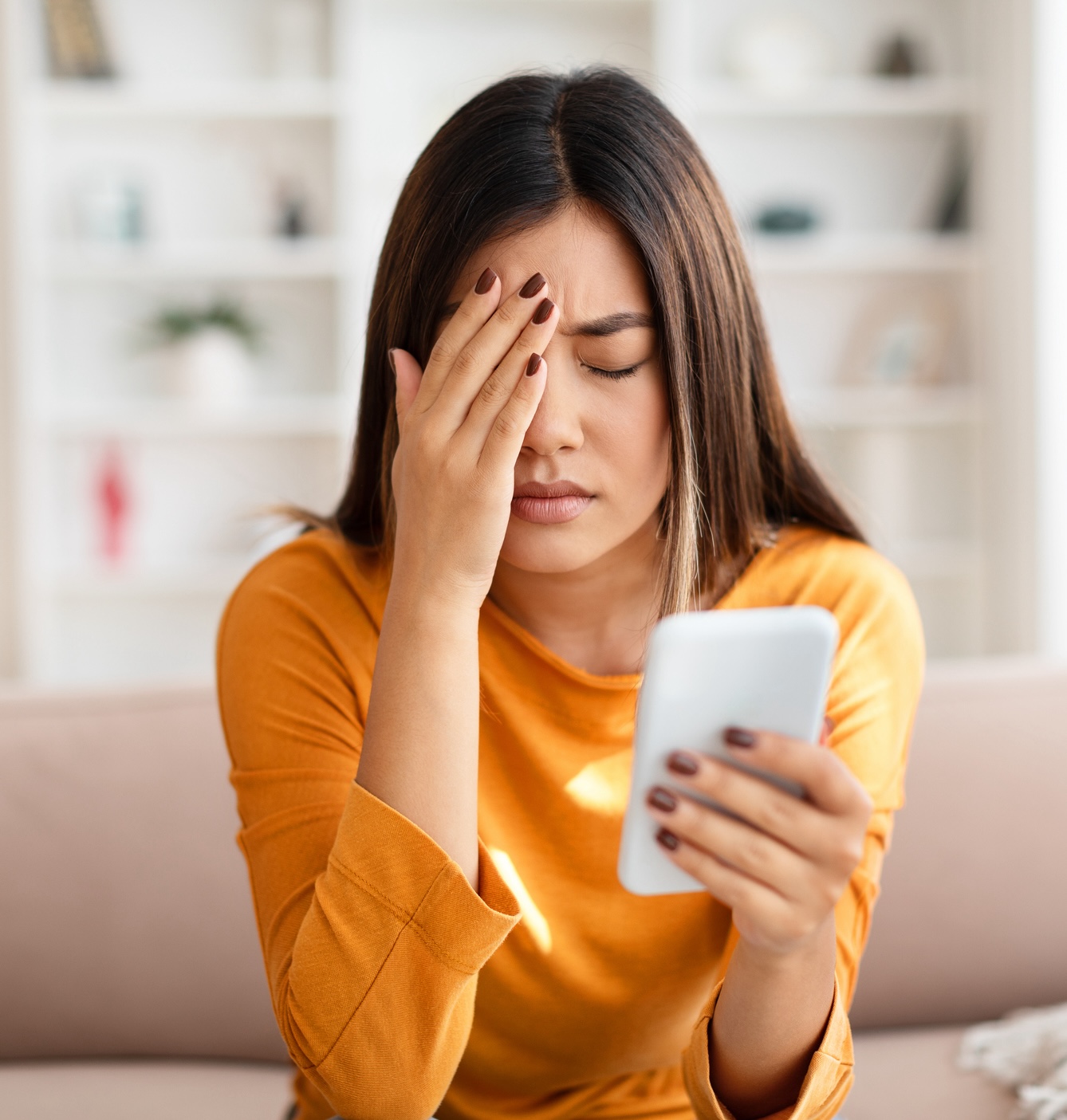 Woman in orange shirt holding phone with eyes closed and hand on forehead, appearing stressed or upset.