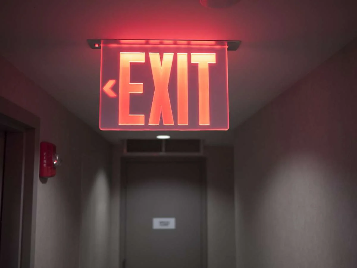 Glowing red exit sign with left arrow hanging from ceiling in a dim hallway.