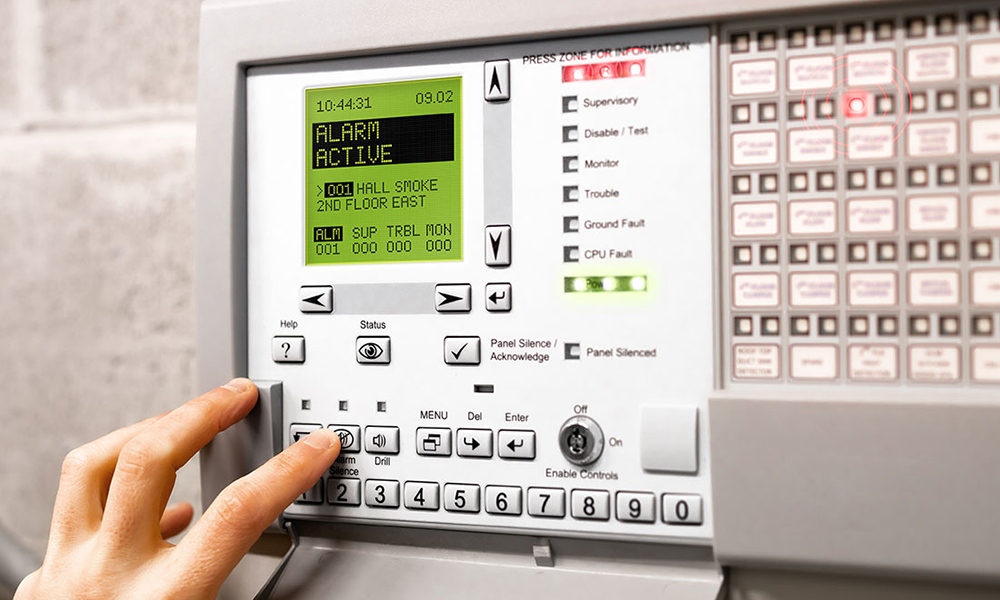 Hand pressing button on fire alarm control panel showing active alarm for hall smoke on 2nd floor east.