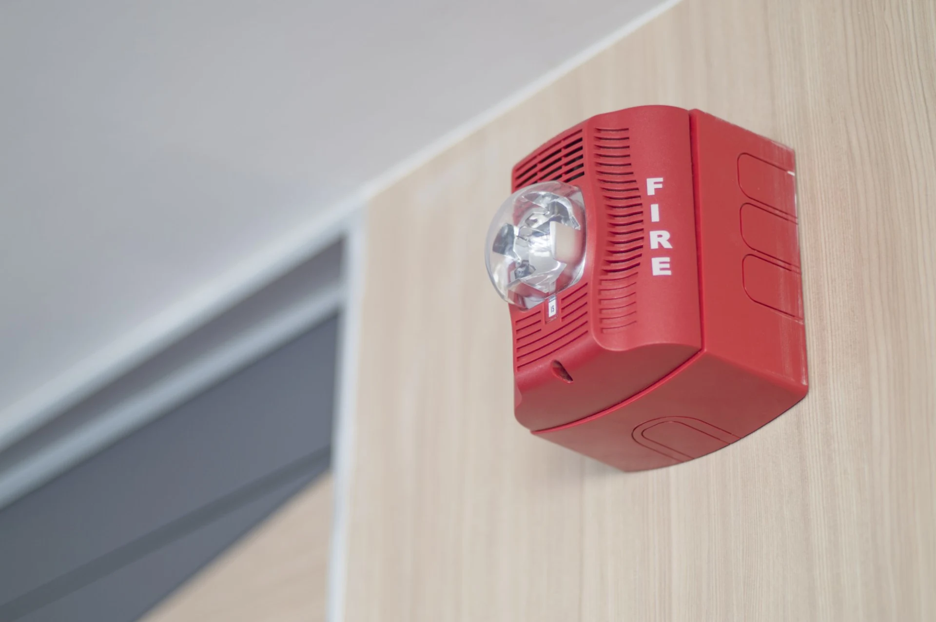 Red fire alarm with clear strobe light mounted on a light wood-paneled wall near ceiling.