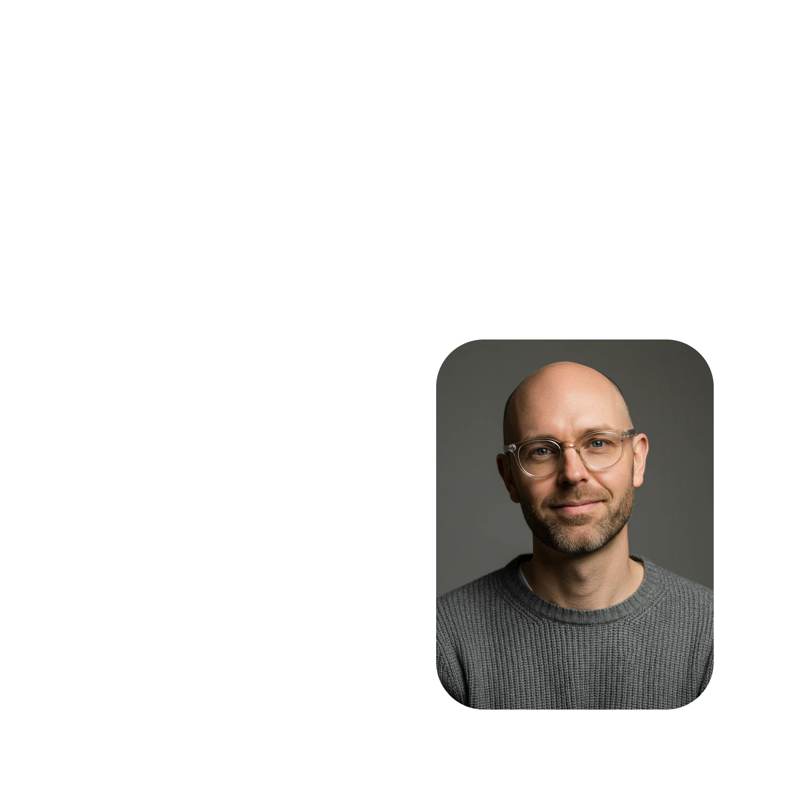Portrait of a smiling bald man with a beard wearing clear glasses and a grey knit sweater against a dark background.