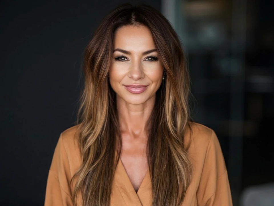 Smiling woman with long brown hair wearing a tan blouse against a dark background.