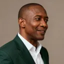 Smiling man in a dark green suit jacket and white shirt against a plain background.