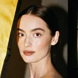 Portrait of a young woman with dark hair, natural makeup, and neutral expression against a blurred background.