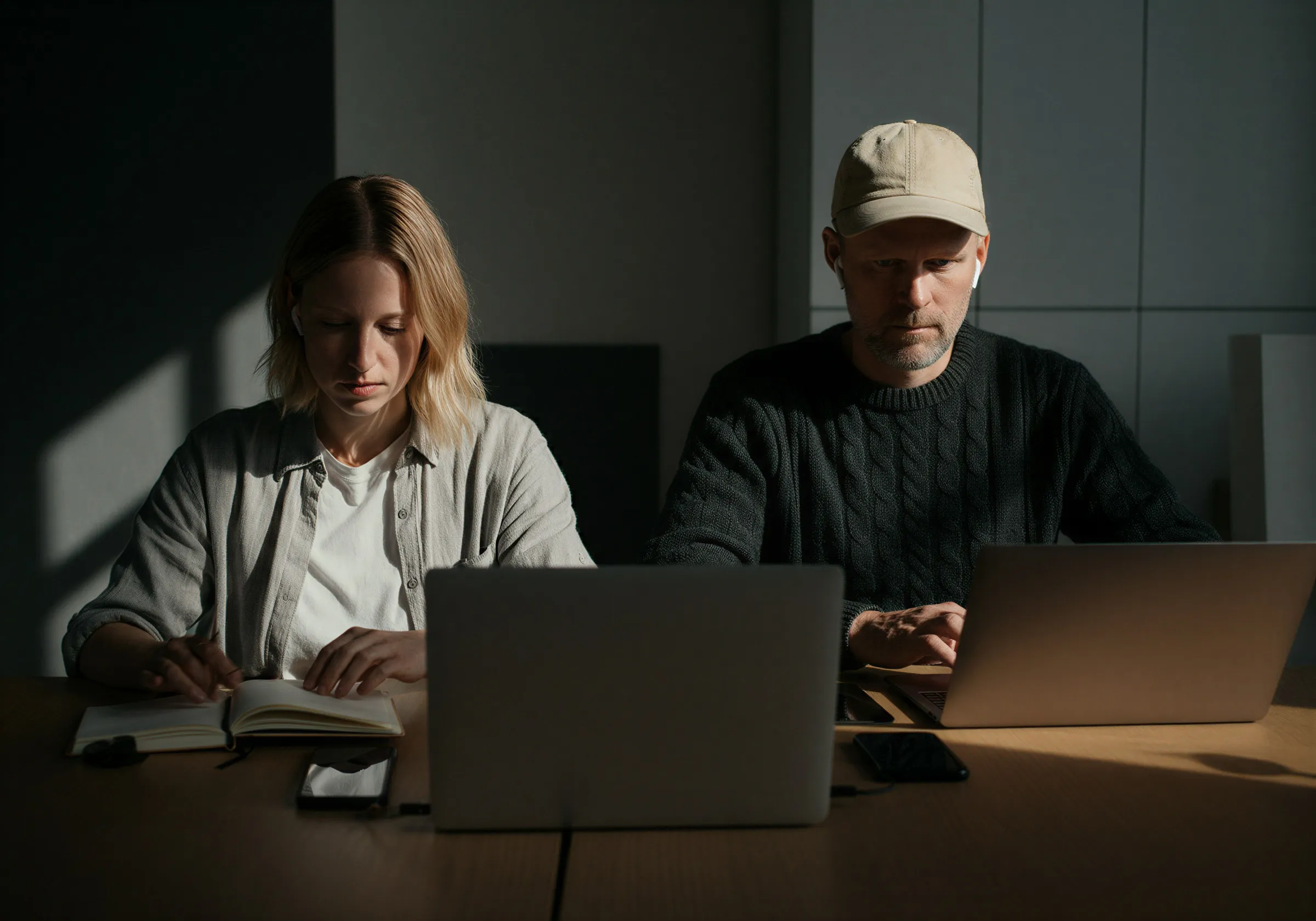 Two people sitting at a table in a dimly lit room, one reading a book and the other working on a laptop.