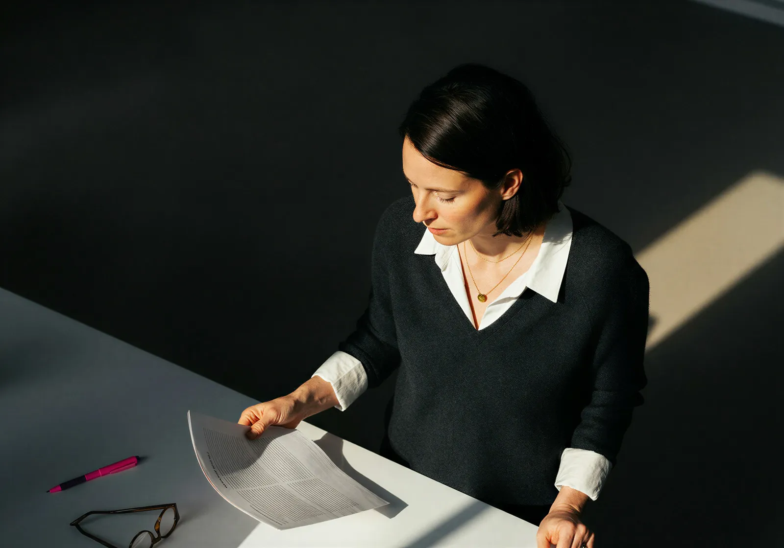 Woman in white shirt and dark sweater reading a document at a white desk with glasses and a pink pen nearby.