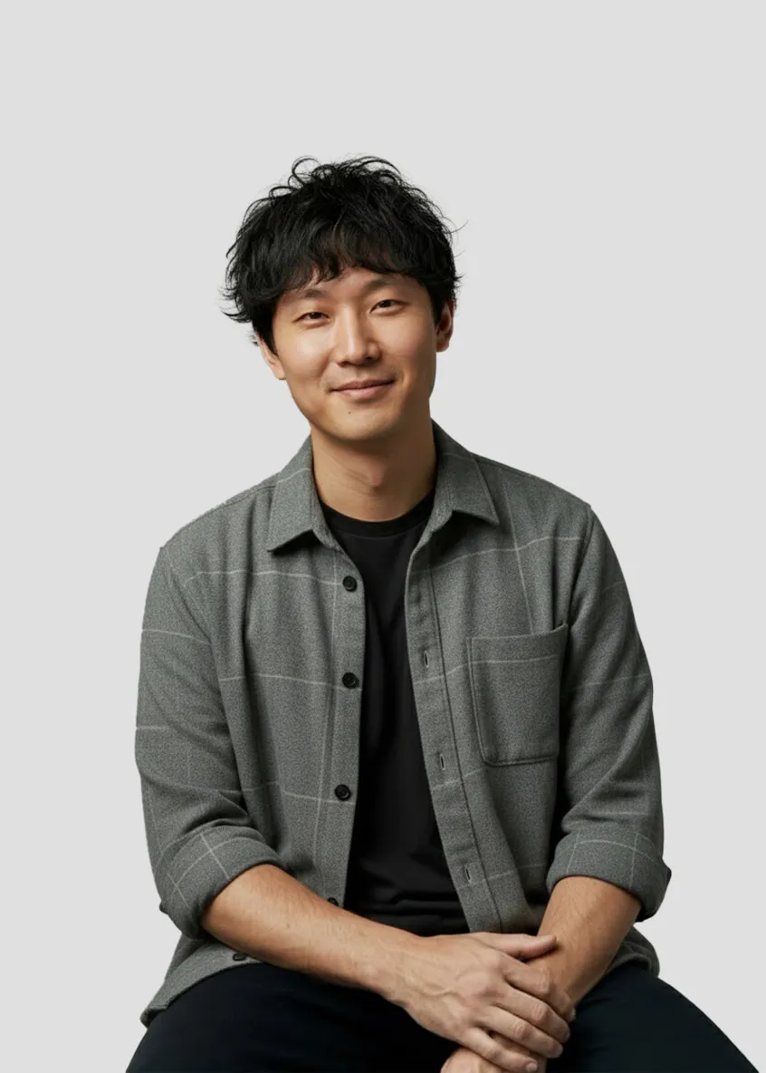 Man with black hair wearing a gray checkered shirt over a black t-shirt, smiling with hands clasped.