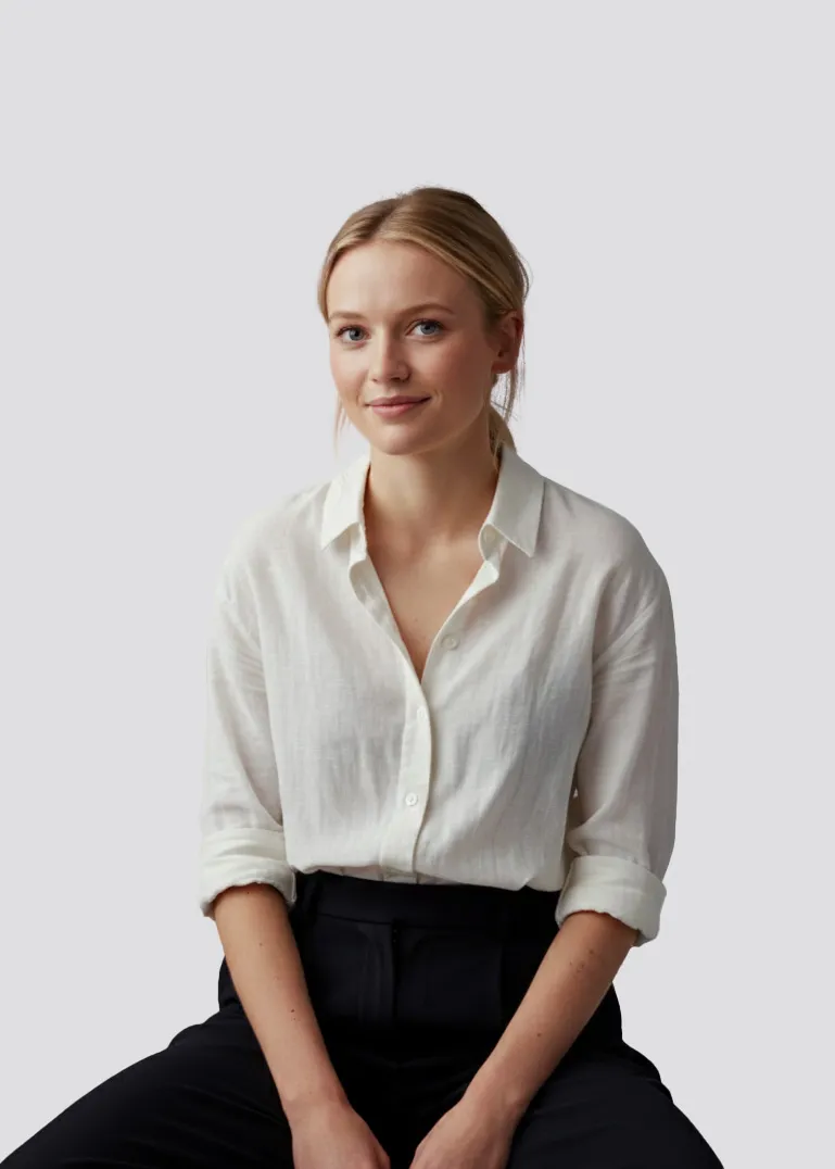 Smiling young woman with blonde hair tied back, wearing a white button-up shirt and black pants, sitting against a plain light gray background.