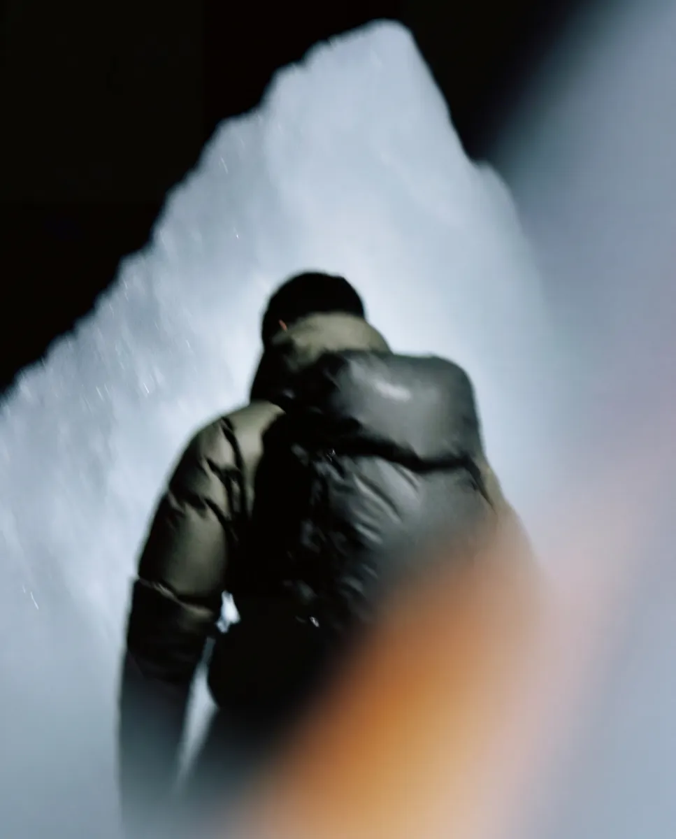 Person wearing a dark jacket and carrying a large backpack inside a dimly lit icy cave.