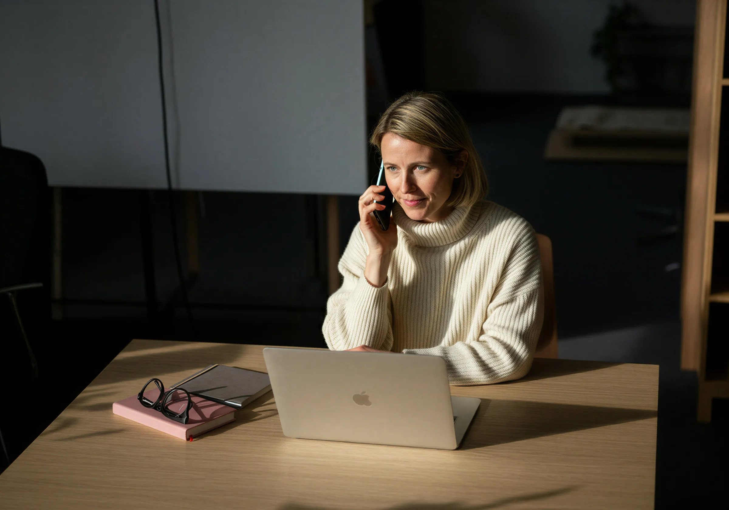 Woman in a white sweater sitting at a desk with a laptop, talking on a smartphone.