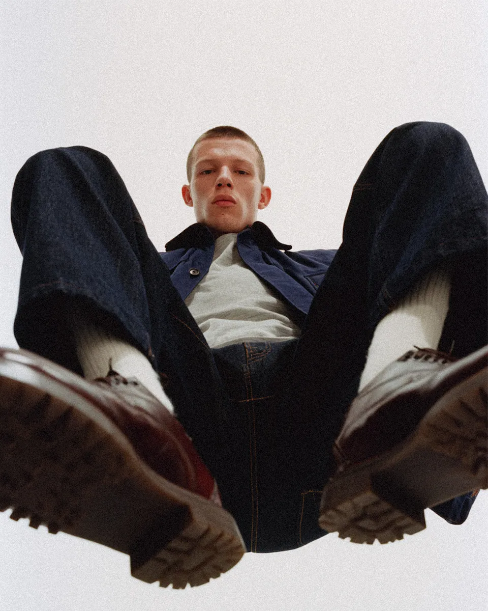 Low-angle view of a man in dark jeans, white socks, brown shoes, and a blue jacket against a plain background.