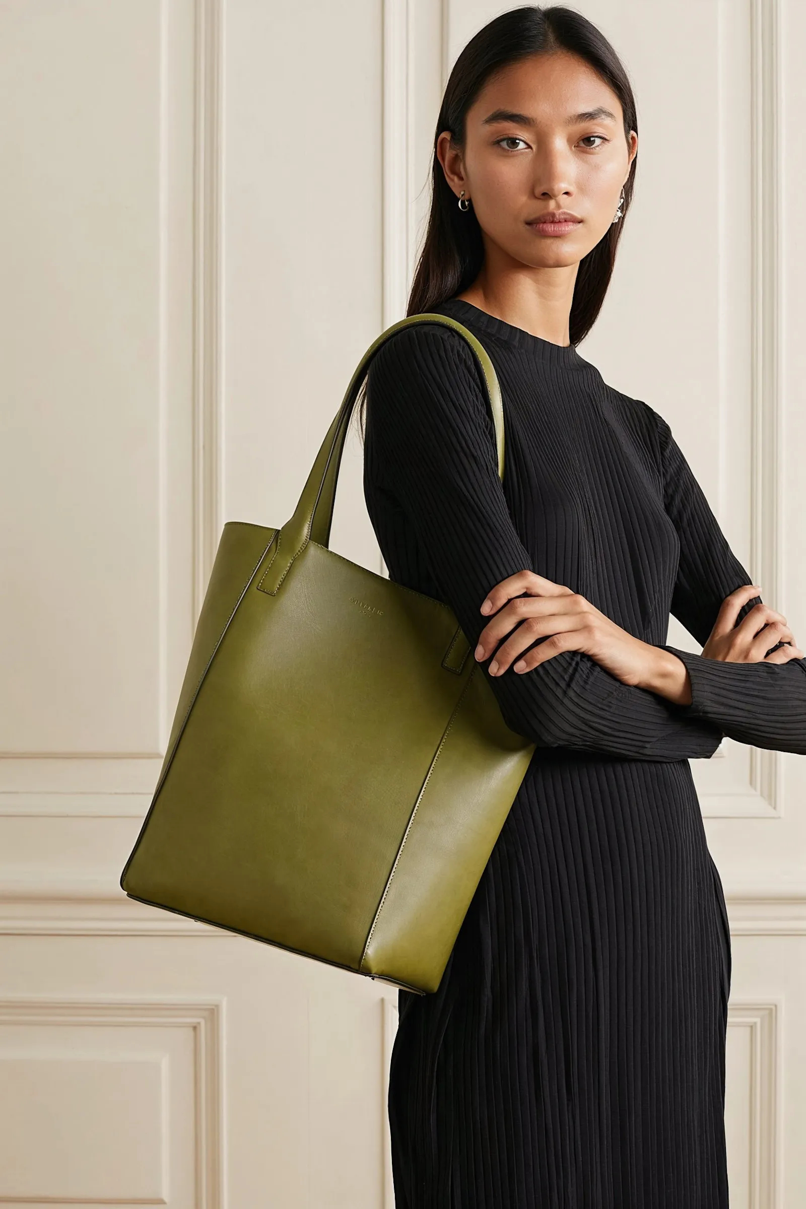 Woman in a black ribbed dress carrying a large olive green leather tote bag on her shoulder.