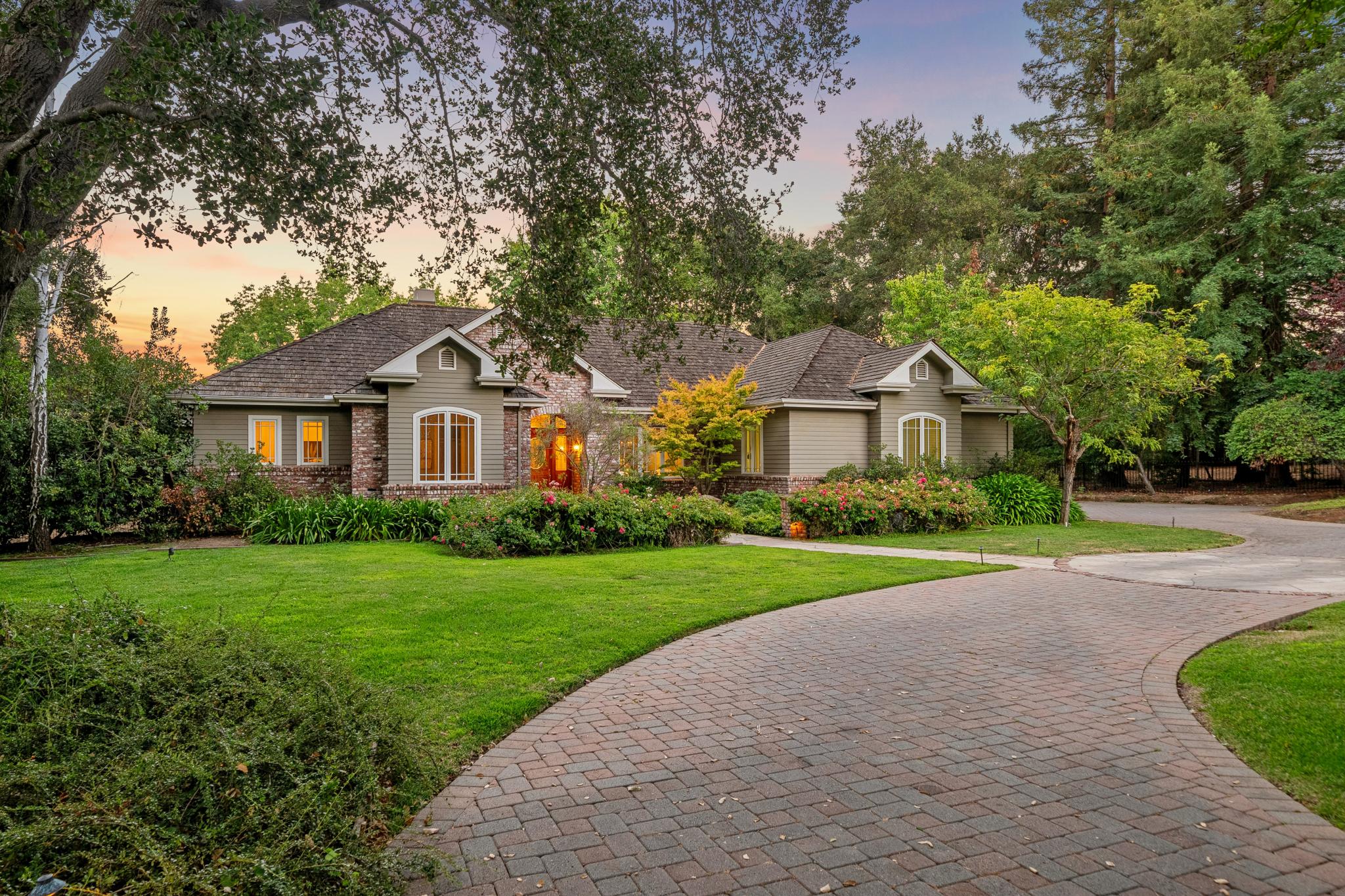 Single-story house with beige siding and brick accents, large arched windows, surrounded by lush green lawn, flowering shrubs, and mature trees at sunset with a curved brick driveway leading to the front.