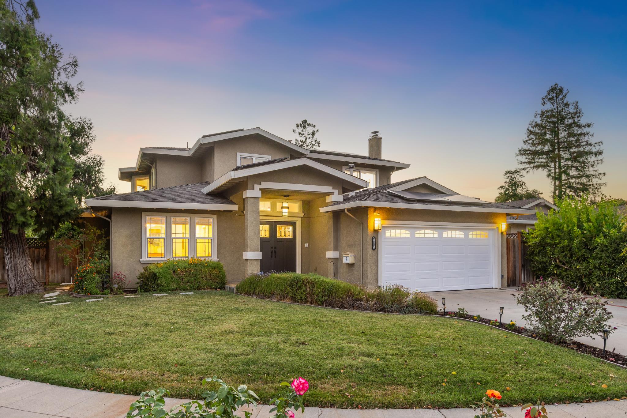 Two-story suburban house with beige exterior, dark double front doors, lit windows, and a white garage door, surrounded by green lawn and trees during dusk.