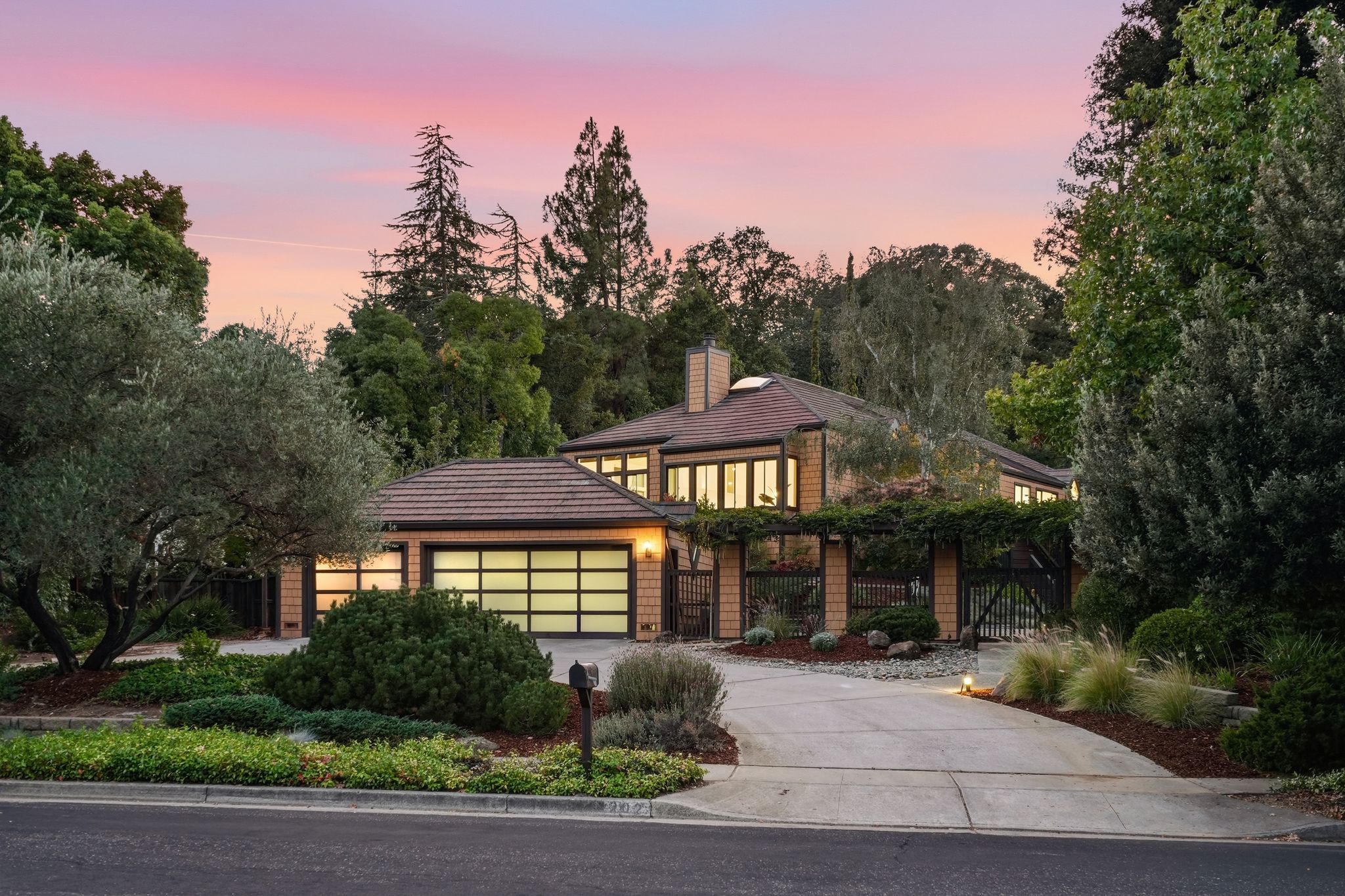 Two-story house with large windows and a glowing garage door, surrounded by lush landscaping and tall trees under a pink and purple twilight sky.