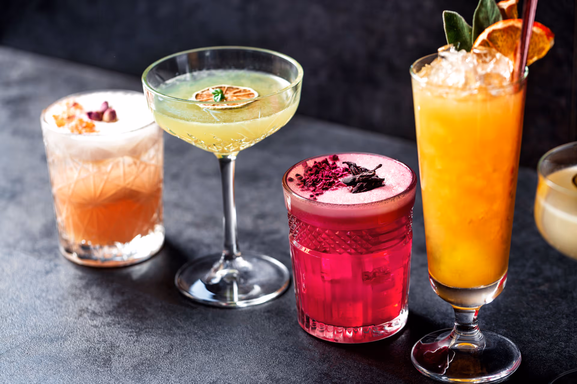 Five colorful cocktails in various glassware, each garnished with herbs or fruit, on a dark surface.