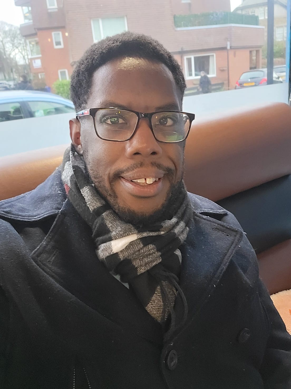 Ricky smiling, wearing glasses, a dark coat and a checked scarf.