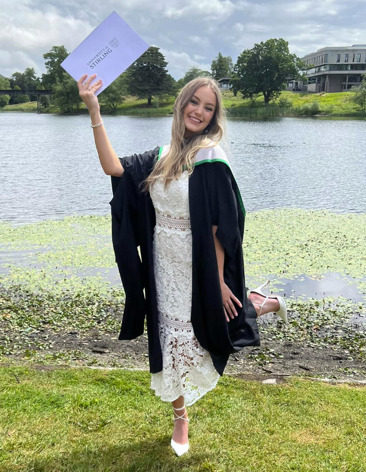 Jodi, in a graduation gown, holding up her certificate beside a lake.