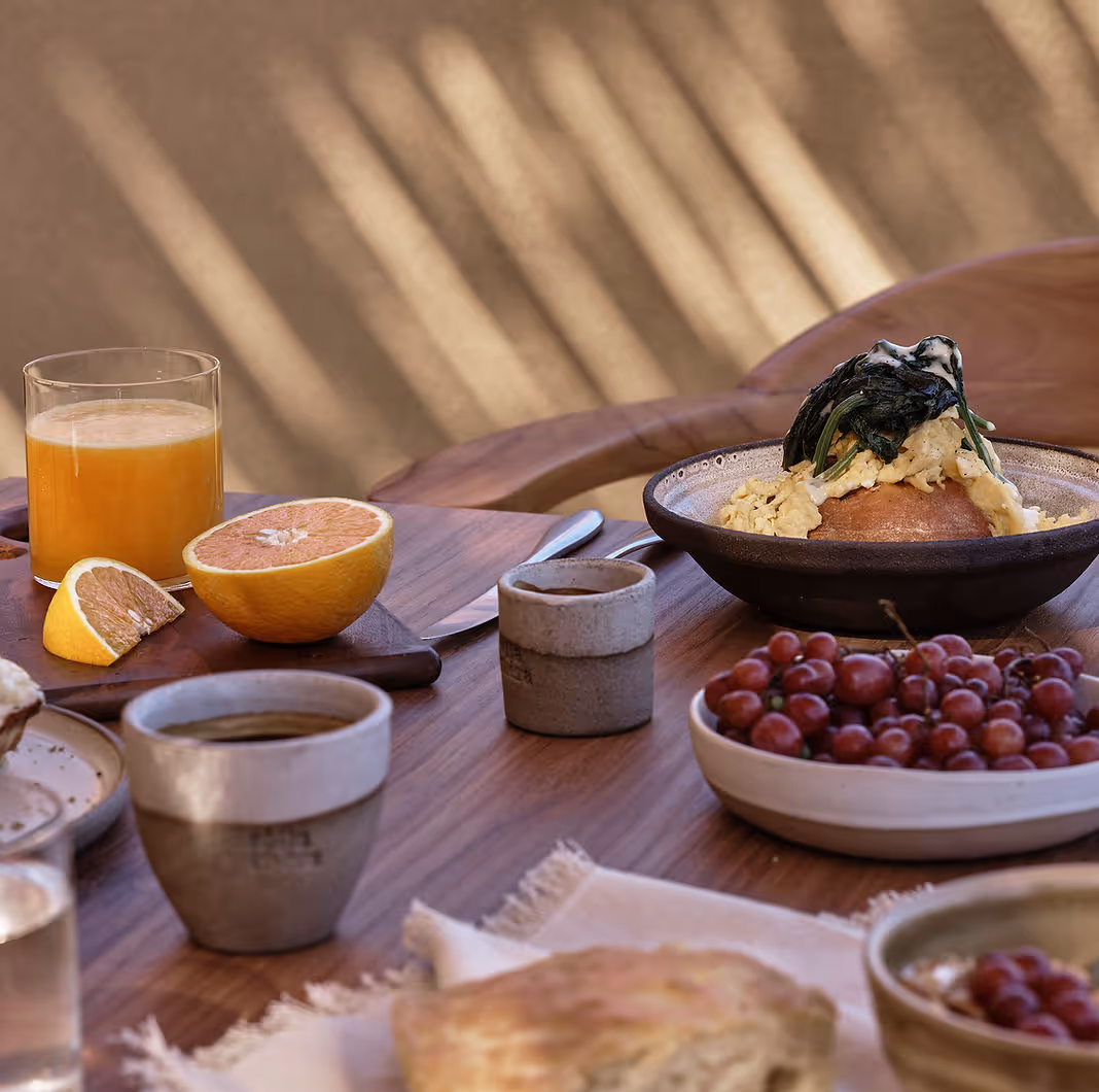 Breakfast table with a glass of orange juice, grapefruit halves, coffee cup, bowl of scrambled eggs on bread with herbs, and a plate of red grapes.