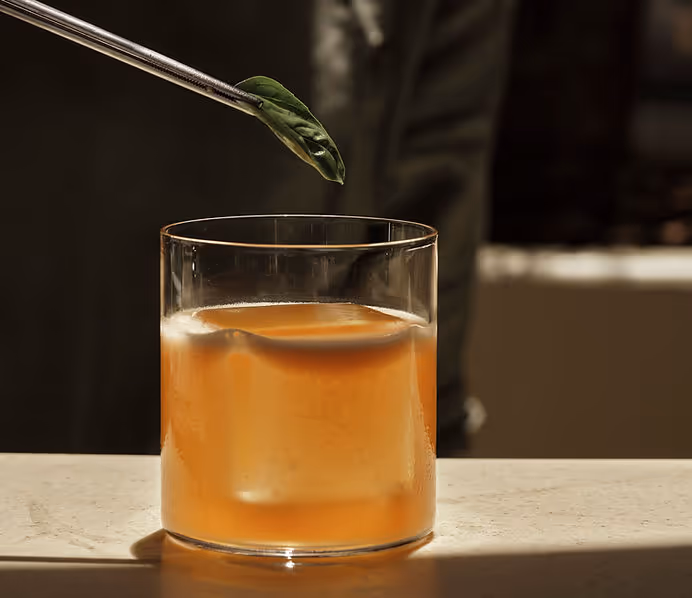 Close-up of a glass with an orange iced drink and a large ice cube, with a pair of tweezers holding a green leaf above it.