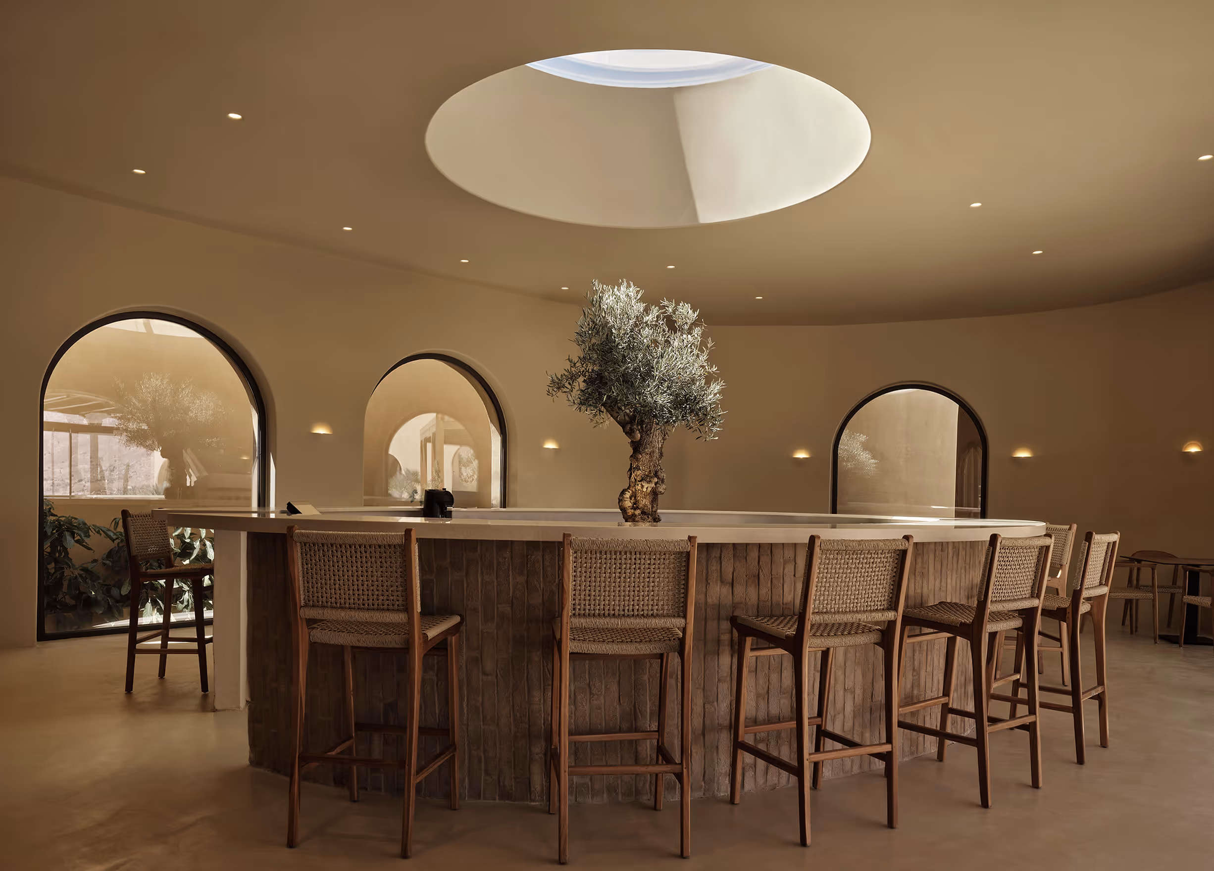 Modern round bar with wooden stools and a tree centerpiece under a circular skylight in a minimalistic beige interior.