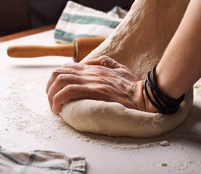 Close-up of hands kneading dough on a floured surface with a rolling pin and kitchen towel in the background.
