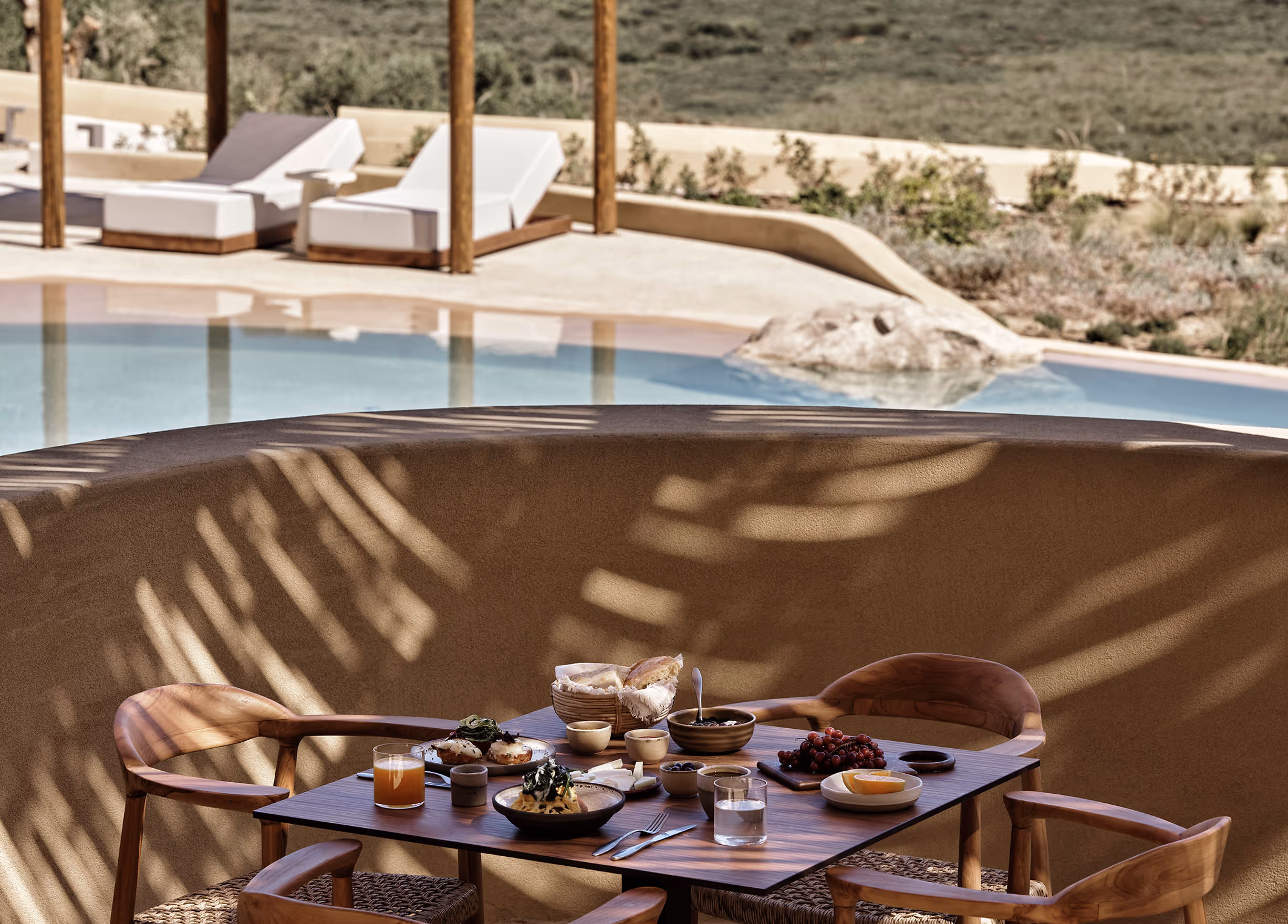 Outdoor wooden table set for breakfast with bread, fruit, juice, and dishes beside a pool and white lounge chairs.