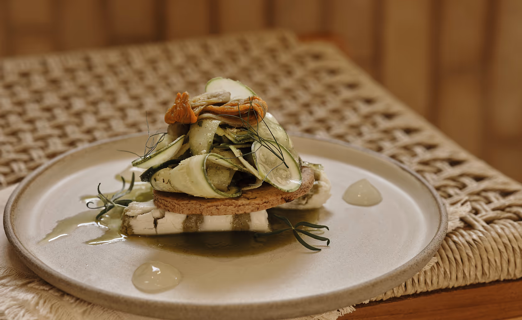 Gourmet dish with thin cucumber slices, edible flowers, and herbs on a round cracker atop a piece of cheese on a beige ceramic plate.