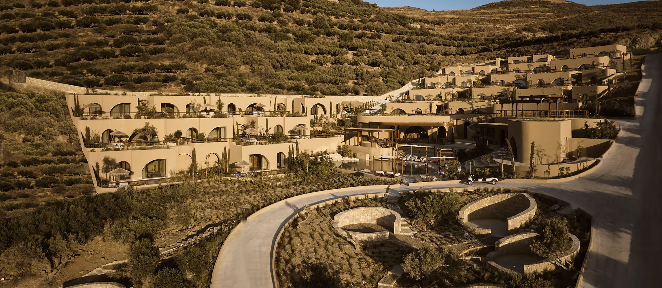 Modern terraced beige hotel complex built into a green hillside with outdoor seating and pathways.