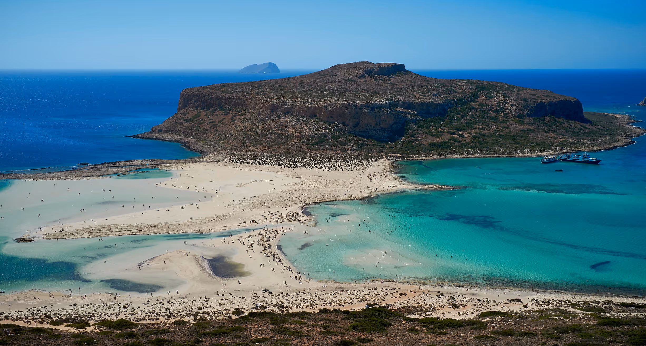 Aerial view of a large rocky island with surrounding turquoise and blue waters and a white sandy beach with people and boats.