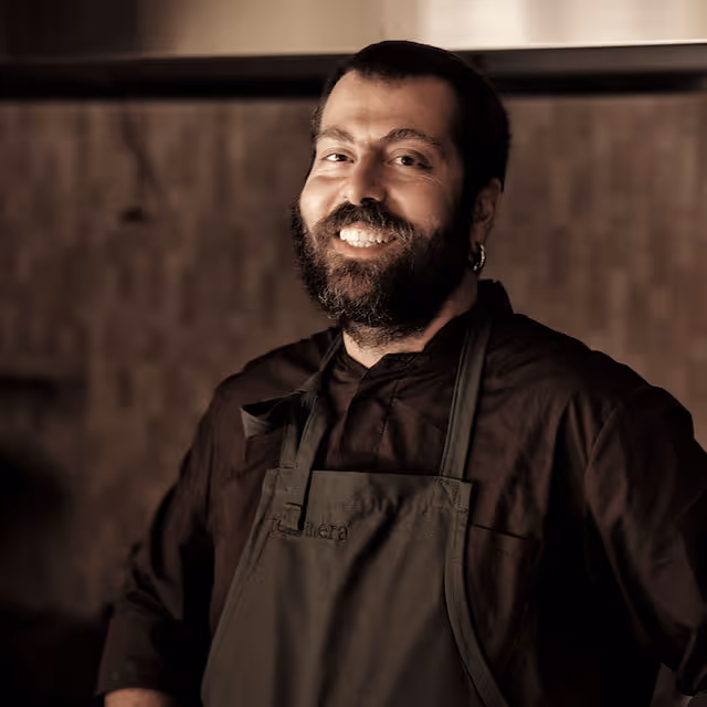 Smiling bearded chef wearing a dark apron and shirt, standing in a warmly lit kitchen.