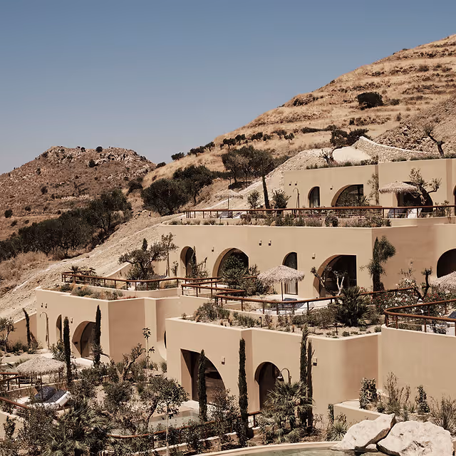 Terraced beige buildings with arched openings built into a hillside with sparse vegetation under a clear sky.