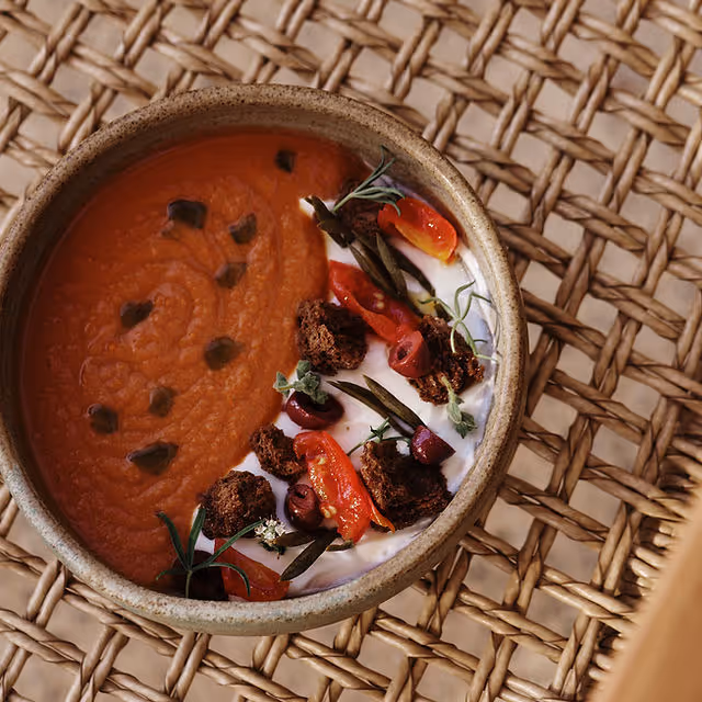 Bowl of creamy orange soup garnished with dollops of white cream, cherry tomatoes, olives, herbs, and brown croutons placed on a woven rattan surface.