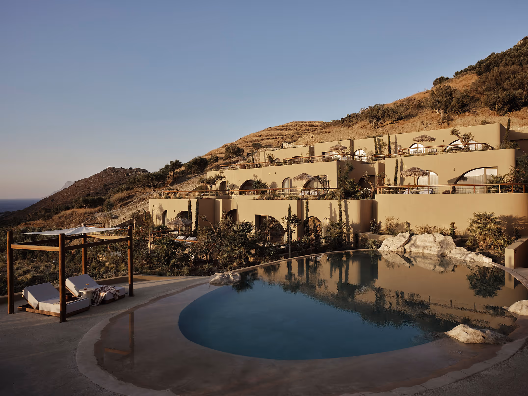 Modern beige terraced buildings on a hillside overlooking a round swimming pool with lounge chairs under a canopy.