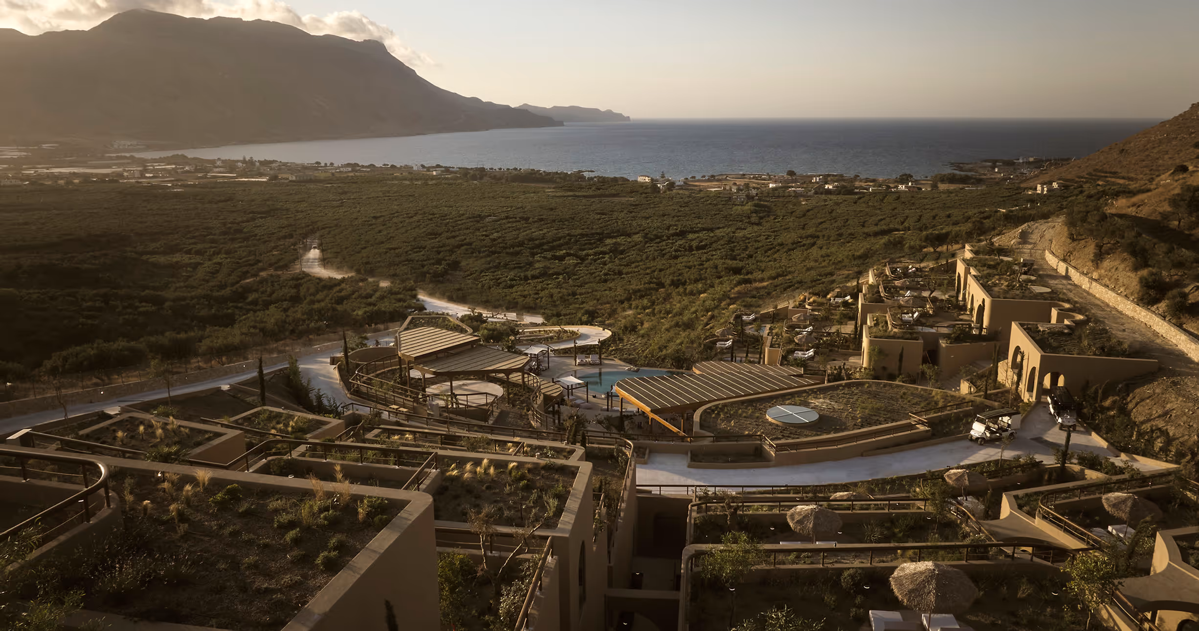Elevated view of a resort with terraced gardens and buildings overlooking green fields and a coastline at sunset.