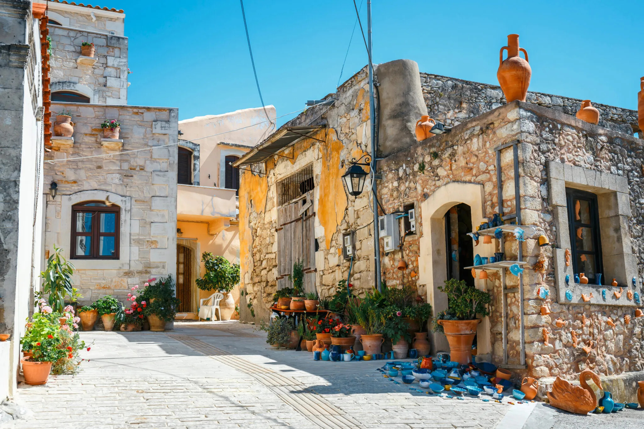 Sunlit narrow stone street in a Mediterranean village with potted plants, flower pots, and blue ceramic pottery displayed outside rustic stone buildings.