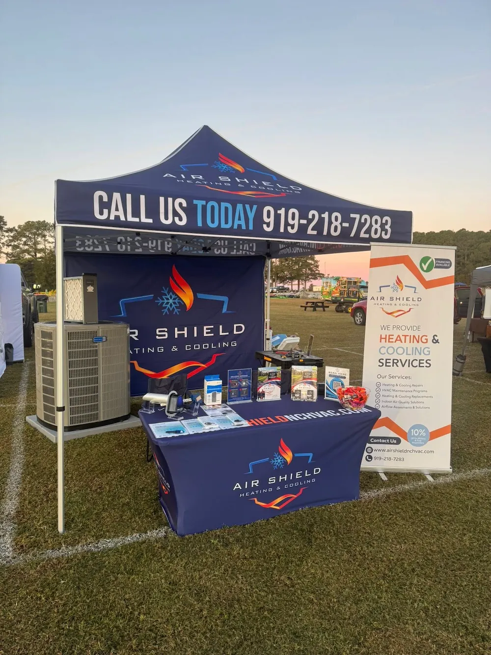 Outdoor booth setup for Air Shield Heating & Cooling with branded tent, table displaying products, air conditioning unit, and a banner listing HVAC services.