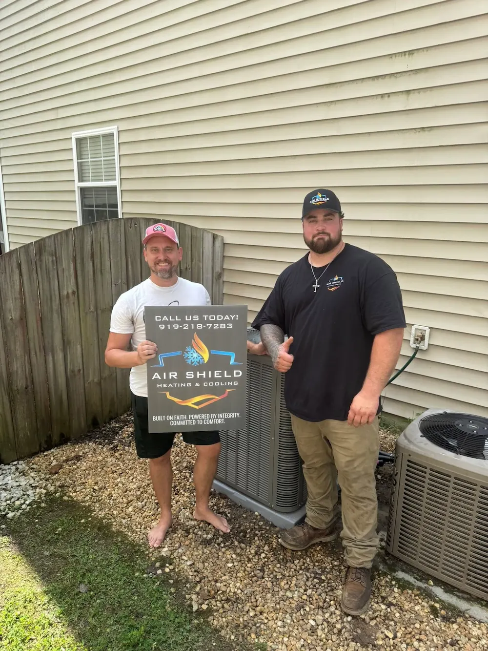 Two men standing outside next to HVAC units, one holding a sign for Air Shield Heating & Cooling with contact number and slogan.