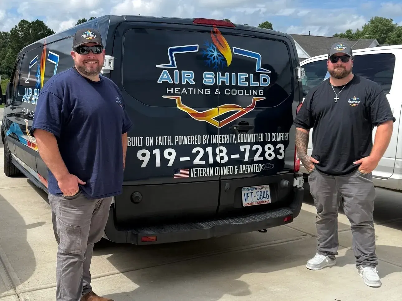 Two men wearing Air Shield Heating & Cooling apparel stand beside a company van with contact number 919-218-7283 and the tagline 'Built on faith, powered by integrity, committed to comfort.'