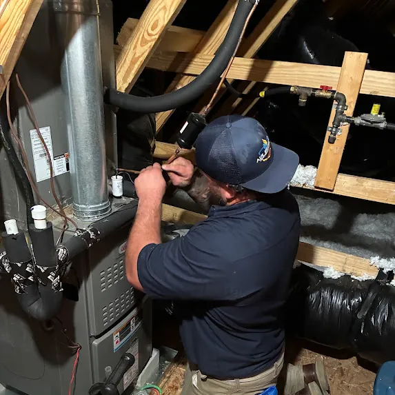 Technician in a navy shirt and cap working on a furnace system in an attic with exposed wooden beams and insulation.