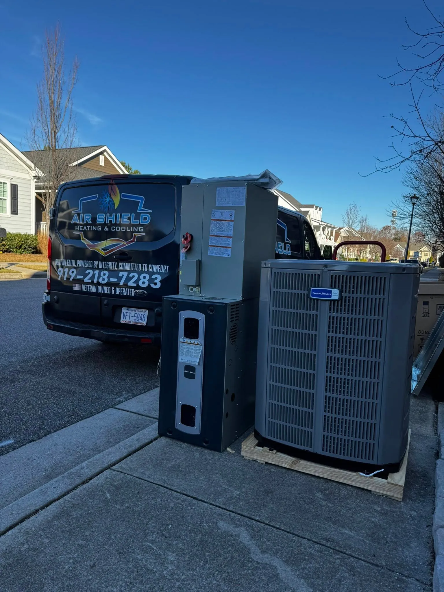 New HVAC equipment on a sidewalk next to a black Air Shield Heating & Cooling service van in a residential neighborhood.