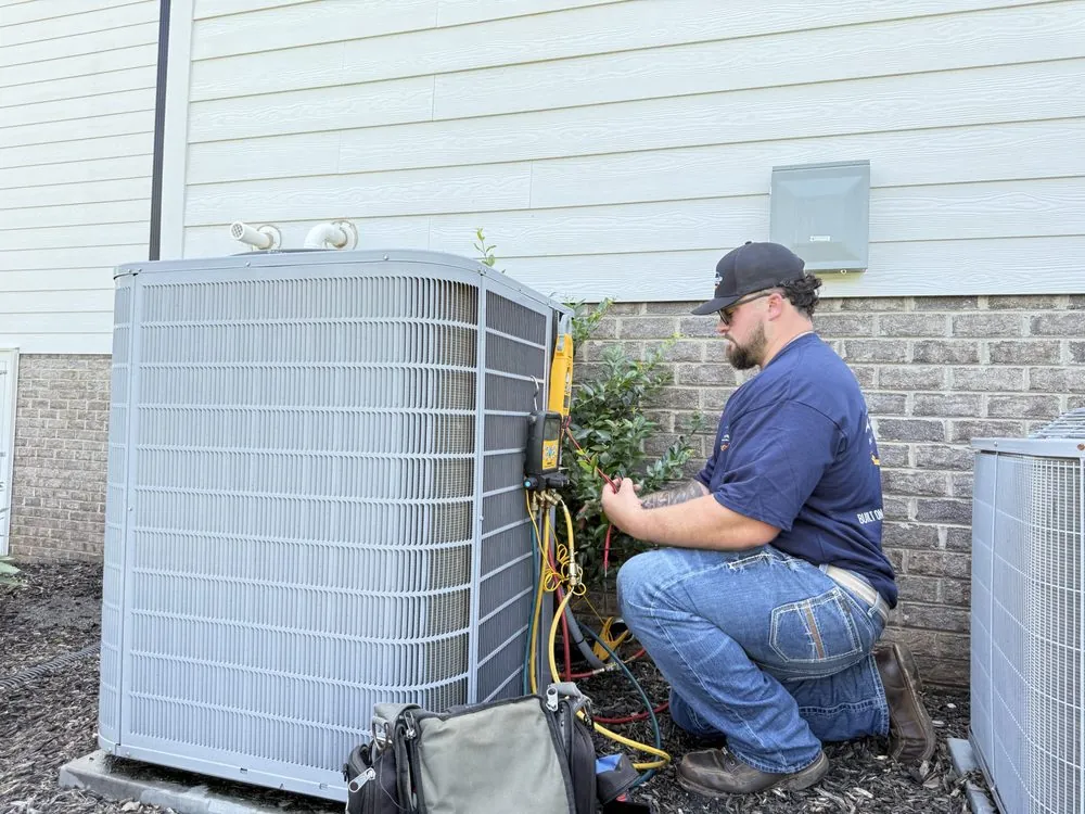 Technician in blue shirt and cap kneeling while checking outdoor air conditioning unit with diagnostic tools.