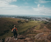 Person hiking on rocky terrain overlooking a green valley and distant city under a partly cloudy sky.