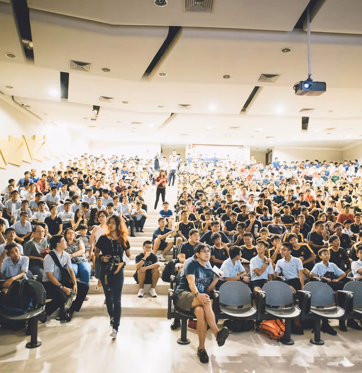 Large lecture hall filled with students, some seated and others standing or walking, under bright ceiling lights.