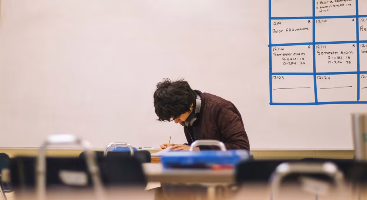 Student sitting alone at a desk in a classroom, writing with a pencil and wearing headphones around the neck.