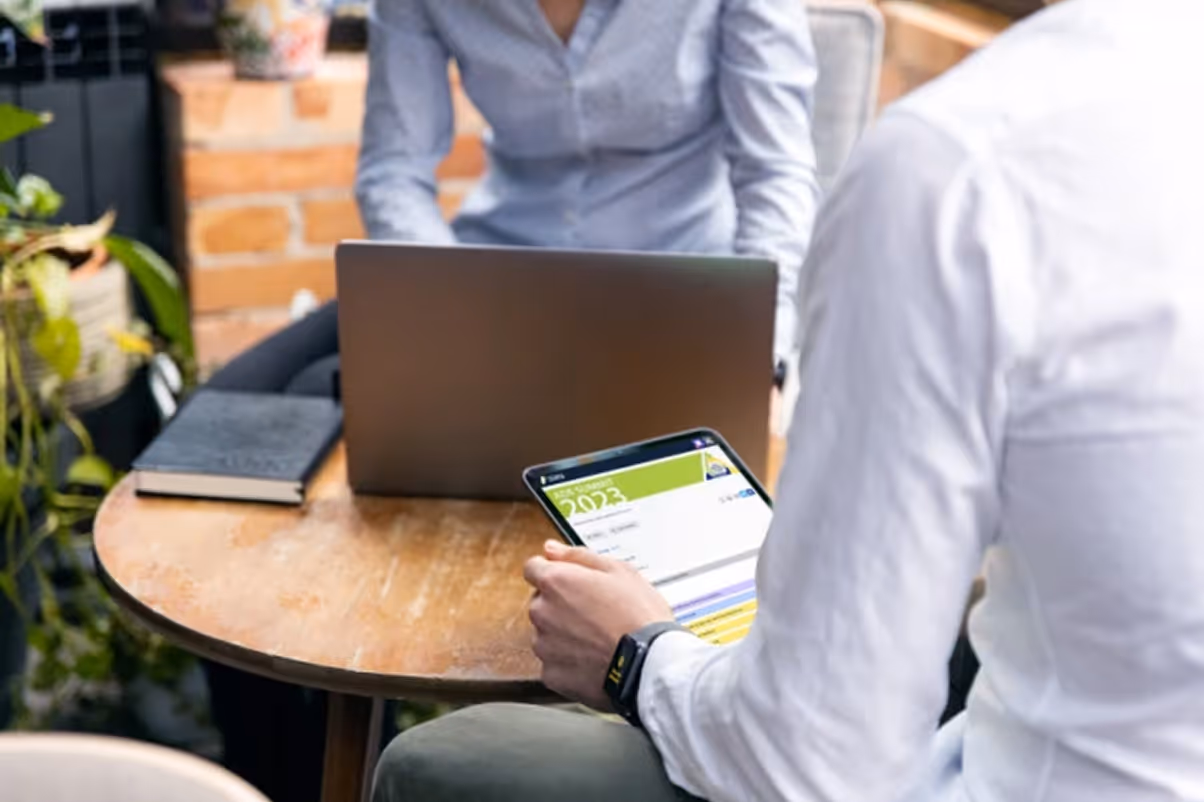 Two people at a round wooden table, one using a laptop and the other holding a tablet displaying a 2023 document.