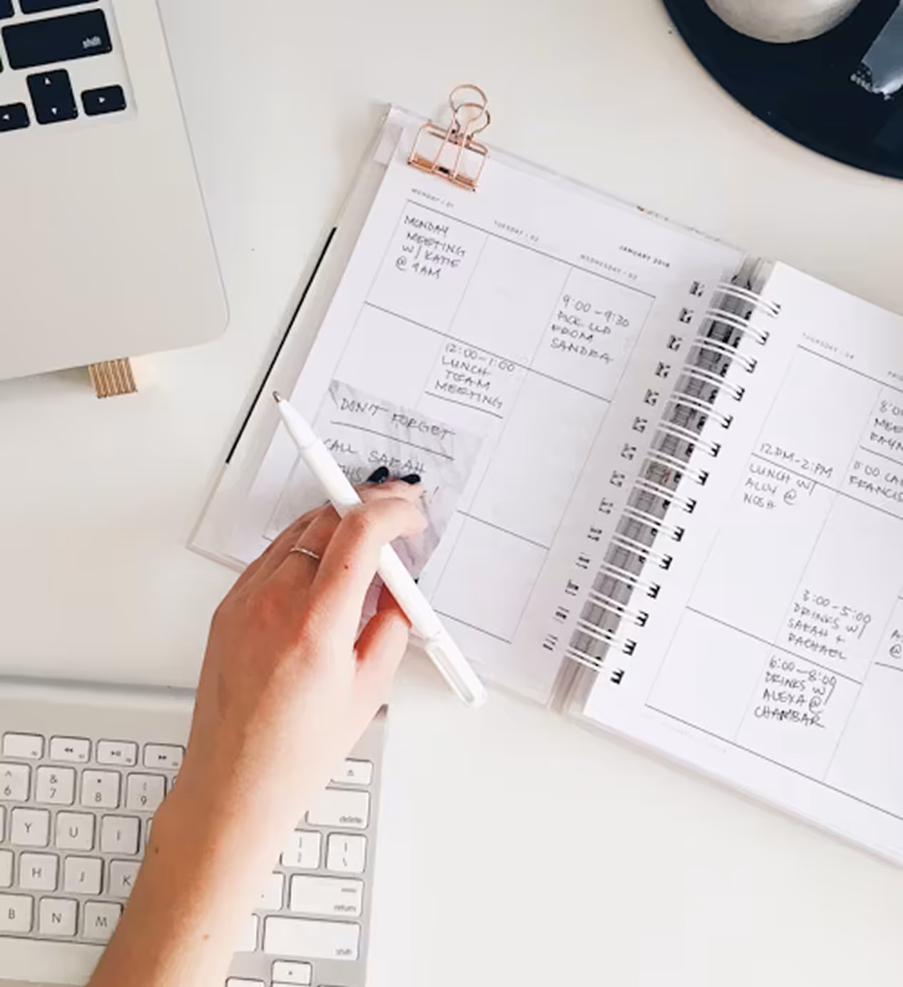Hand holding a white pen over an open spiral notebook calendar with handwritten notes beside a keyboard and laptop on a white desk.